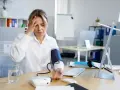 Kobieta z bólem głowy mierzy ciśnienie, podejrzewając hipotonię. Na biurku aparat, szklanka wody i komputer.