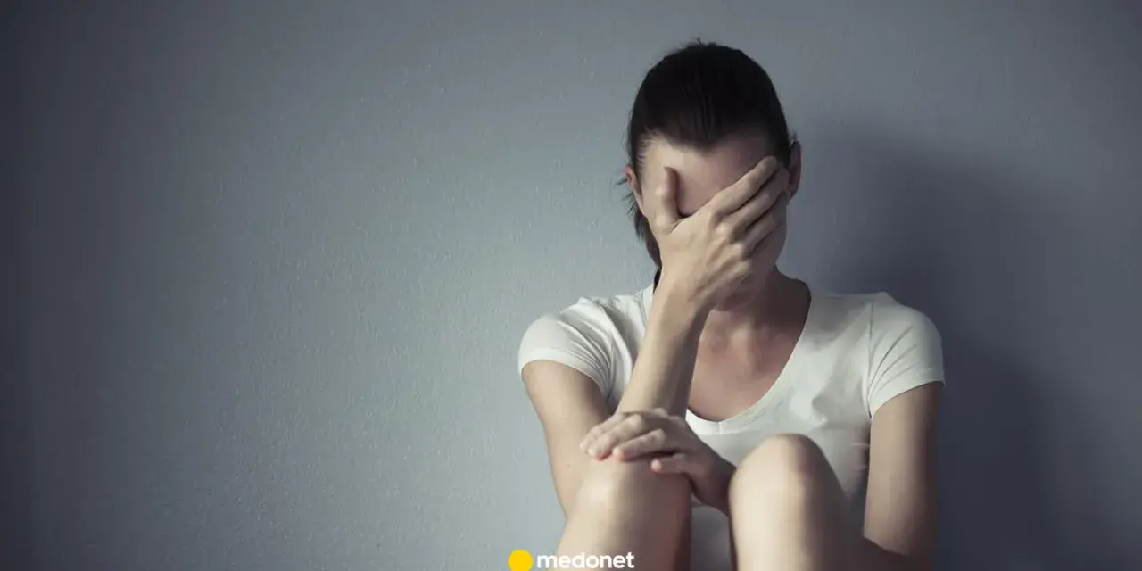 Kobieta w białej koszulce zakrywa twarz dłońmi, siedząc przy szarej ścianie. Może to być symbol smutku lub problemów, z którymi zmaga się, jak np. depresja, którą czasem leczy się lekiem lerivon.