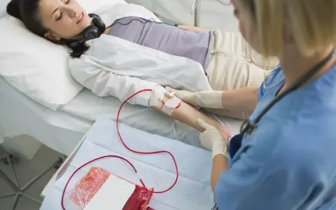 Kobieta leży w łóżku szpitalnym, słuchając muzyki przez słuchawki. Pielęgniarka wykonuje przetaczanie krwi, podłączając worek z krwią do jej ręki.