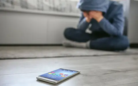 Osoba w kapturze siedzi na podłodze, ukrywając twarz w dłoniach. Obok leży telefon, symbolizujący izolację i dystymię.