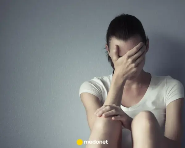 Kobieta w białej koszulce zakrywa twarz dłońmi, siedząc przy szarej ścianie. Może to być symbol smutku lub problemów, z którymi zmaga się, jak np. depresja, którą czasem leczy się lekiem lerivon.