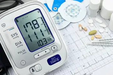 Cyfrowy ciśnieniomierz pokazuje wysokie ciśnienie 178/103. Obok leżą tabletki, elektrody EKG i papier z zapisem EKG, sugerując monitorowanie holterem ciśnieniowym.